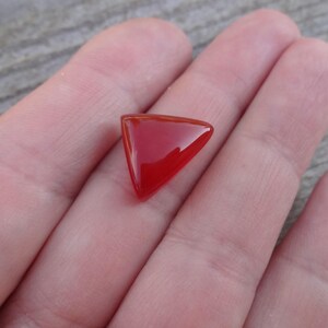 Puede incluir: Una gema roja triangular y pulida. La gema es de un rojo vibrante y brillante con una superficie lisa. La forma es un tri&aacute;ngulo perfecto con esquinas redondeadas. La piedra preciosa se sostiene en una mano, con una superficie de madera en el fondo.