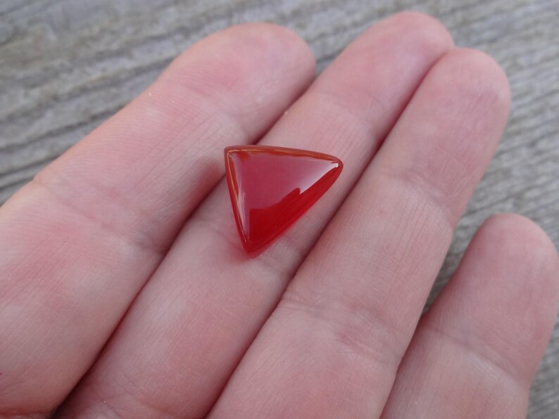 Puede incluir: Una gema roja triangular y pulida. La gema es de un rojo vibrante y brillante con una superficie lisa. La forma es un tri&aacute;ngulo perfecto con esquinas redondeadas. La piedra preciosa se sostiene en una mano, con una superficie de madera en el fondo.