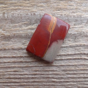 May include: A red, white, and orange jasper cabochon with a smooth, polished surface. The cabochon is rectangular in shape and has a unique, swirling pattern.