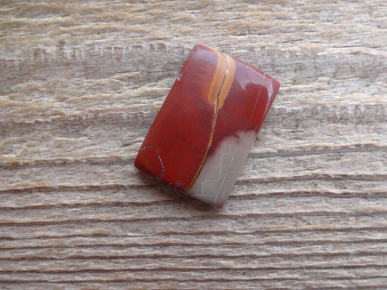 May include: A red, white, and orange jasper cabochon with a smooth, polished surface. The cabochon is rectangular in shape and has a unique, swirling pattern.