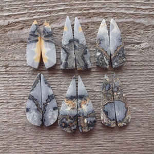 Puede incluir: Seis pares de colgantes de piedras preciosas pulidas de varias formas. Las piedras presentan patrones grises, negros y beige. Los colgantes se exhiben sobre una superficie de madera, mostrando sus colores y texturas naturales. Ideal para la creaci&oacute;n de joyas.