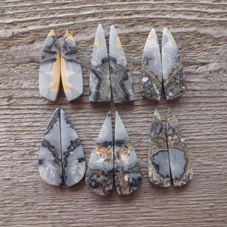 Puede incluir: Seis pares de colgantes de piedras preciosas pulidas de varias formas. Las piedras presentan patrones grises, negros y beige. Los colgantes se exhiben sobre una superficie de madera, mostrando sus colores y texturas naturales. Ideal para la creaci&oacute;n de joyas.