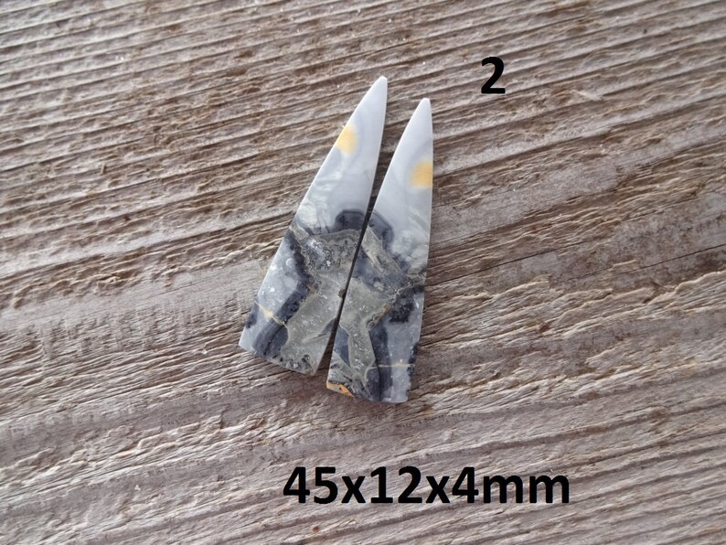 Puede incluir: Dos piedras triangulares pulidas con un patr&oacute;n veteado gris, negro y amarillo. Las piedras est&aacute;n sobre un fondo de madera envejecida. El texto "2" est&aacute; encima de las piedras y "45x12x4mm" debajo.