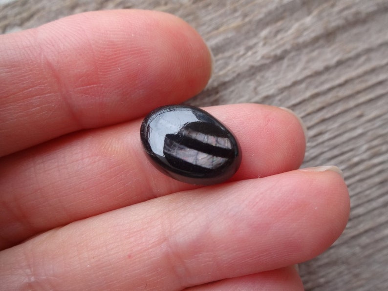 May include: A black oval cabochon gemstone with iridescent stripes. The gemstone is polished and has a smooth, shiny surface.