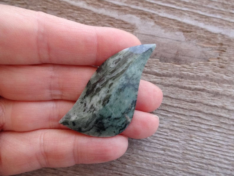 Puede incluir: Una piedra verde y negra con vetas, con una punta puntiaguda, con forma de hoja. La piedra es lisa y pulida.
