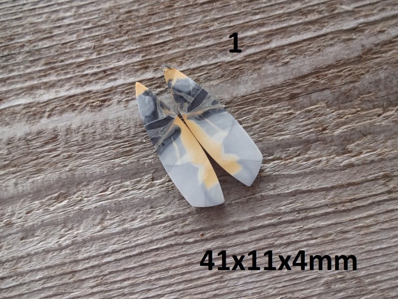 Puede incluir: Dos colgantes alargados de piedra pulida con un patr&oacute;n gris claro, negro y amarillo. Los colgantes est&aacute;n etiquetados con el n&uacute;mero "1" y miden 41x11x4mm.