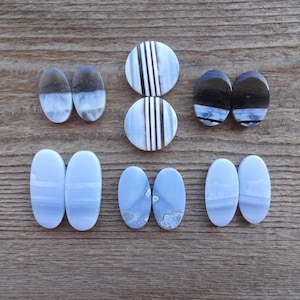 May include: A collection of oval and round cabochon gemstones in shades of blue, white, and black. The gemstones are arranged on a wooden surface, showcasing their unique patterns and colors. The collection includes various sizes and designs.