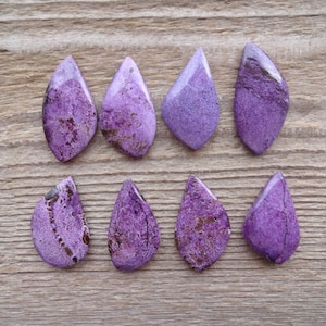 May include: Eight purple and brown gemstone cabochons in a teardrop shape. The cabochons are arranged in two rows of four on a wooden surface.
