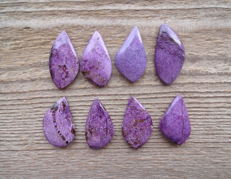 May include: Eight purple and brown gemstone cabochons in a teardrop shape. The cabochons are arranged in two rows of four on a wooden surface.