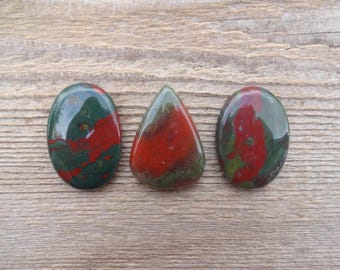 May include: Three polished gemstones with a mix of red and green colors. The gemstones are oval and teardrop shaped. The stones are displayed on a wooden surface.