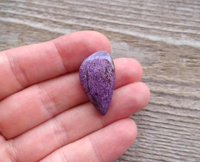 May include: A purple and silver gemstone cabochon with a teardrop shape. The gemstone has a unique pattern of veins and inclusions.