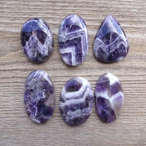 May include: Six polished amethyst gemstones in oval and teardrop shapes. The stones display a mix of deep purple and white banding patterns. The gemstones are arranged on a weathered, light brown wooden surface.