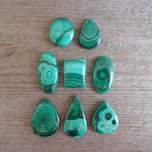 K&ouml;nnte beinhalten: Eine Sammlung von acht gr&uuml;nen Malachit-Cabochons in verschiedenen Formen und Gr&ouml;&szlig;en. Die Cabochons sind in zwei Reihen auf einer Holzoberfl&auml;che angeordnet.