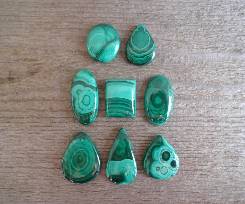 K&ouml;nnte beinhalten: Eine Sammlung von acht gr&uuml;nen Malachit-Cabochons in verschiedenen Formen und Gr&ouml;&szlig;en. Die Cabochons sind in zwei Reihen auf einer Holzoberfl&auml;che angeordnet.