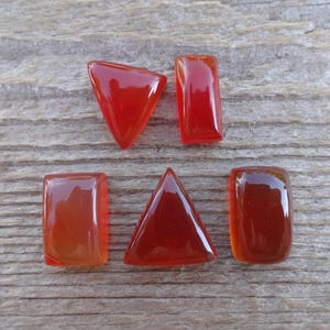 Puede incluir: Seis gemas pulidas, transl&uacute;cidas, de color rojo-naranja, en varias formas: un tri&aacute;ngulo, un trapecio y rect&aacute;ngulos. Las piedras est&aacute;n dispuestas sobre una superficie de madera gris envejecida, mostrando su acabado brillante y rico color.
