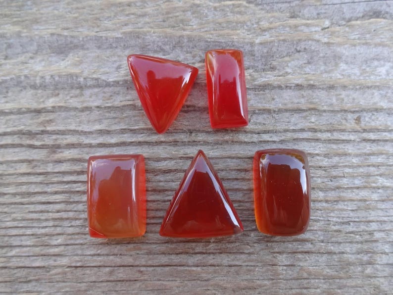 Puede incluir: Seis gemas pulidas, transl&uacute;cidas, de color rojo-naranja, en varias formas: un tri&aacute;ngulo, un trapecio y rect&aacute;ngulos. Las piedras est&aacute;n dispuestas sobre una superficie de madera gris envejecida, mostrando su acabado brillante y rico color.