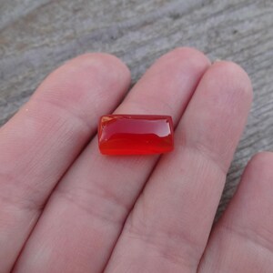 Puede incluir: Una gema roja, rectangular y pulida. La gema se sostiene en una mano, mostrando su color vibrante y superficie lisa. La forma de la piedra es un rect&aacute;ngulo simple, con bordes redondeados.