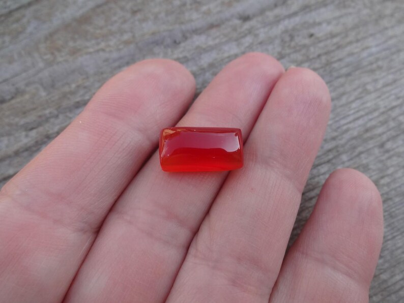 Puede incluir: Una gema roja, rectangular y pulida. La gema se sostiene en una mano, mostrando su color vibrante y superficie lisa. La forma de la piedra es un rect&aacute;ngulo simple, con bordes redondeados.