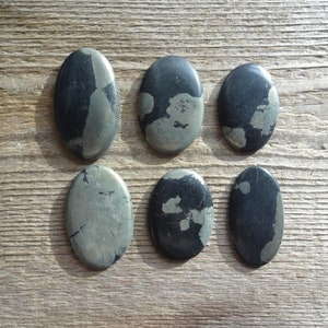 May include: Six oval-shaped pyrite stones with a metallic sheen, arranged on a wooden surface. The stones display a mix of dark gray and silver-gold colors, with a natural, textured appearance. The stones are smooth and polished.