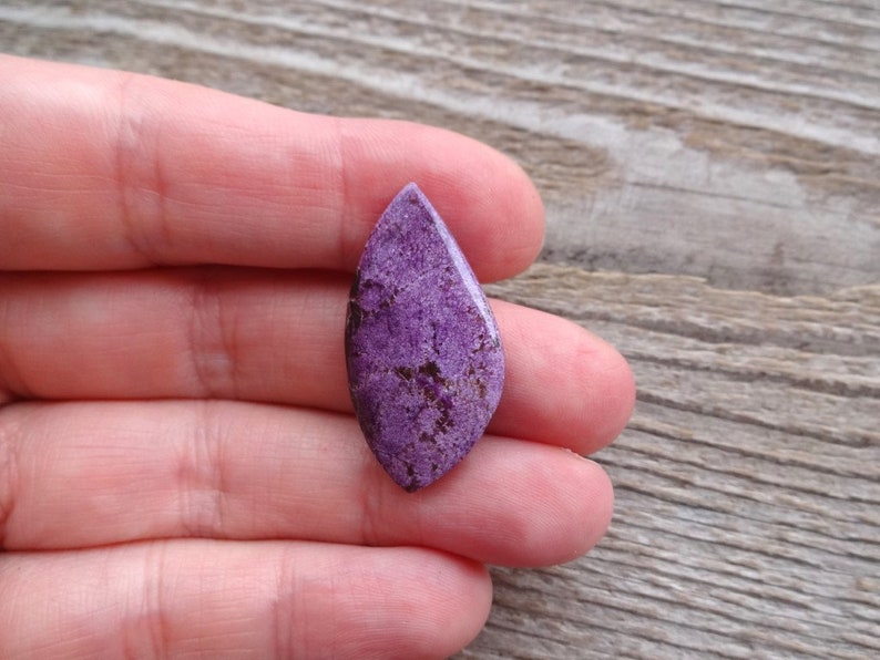 May include: A purple and brown gemstone cabochon with a teardrop shape. The gemstone is set in a silver bezel.
