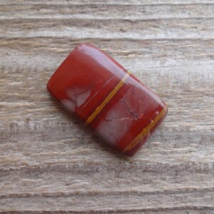 May include: A red jasper cabochon with thin, gold-colored veins. The stone is rectangular and has a smooth, polished surface.