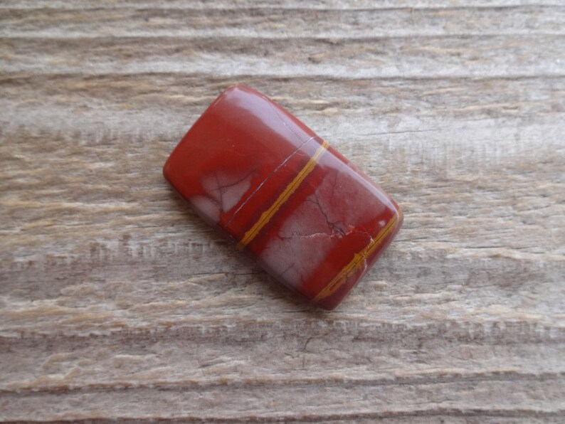 May include: A red jasper cabochon with thin, gold-colored veins. The stone is rectangular and has a smooth, polished surface.