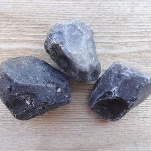 May include: Three dark gray, irregularly shaped stones with a rough texture. The stones are arranged on a light brown, wood-grain surface. The stones appear to be natural and unpolished.