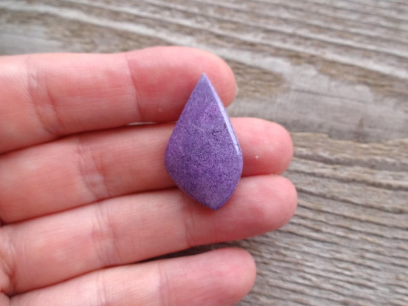 May include: A purple, teardrop-shaped cabochon gemstone with a speckled texture.