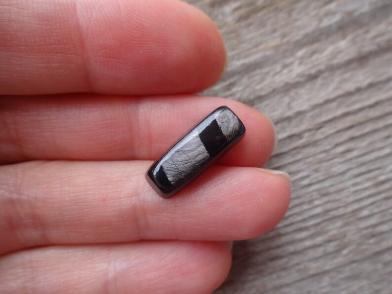 May include: A black and silver rectangular cabochon with a striated pattern. The cabochon is approximately 1 inch long.