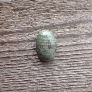 K&ouml;nnte beinhalten: Ein gr&uuml;ner ovaler Cabochon-Edelstein mit einem einzigartigen Muster aus wei&szlig;en und grauen Streifen. Der Stein ist vor einem rustikalen Holzhintergrund platziert.