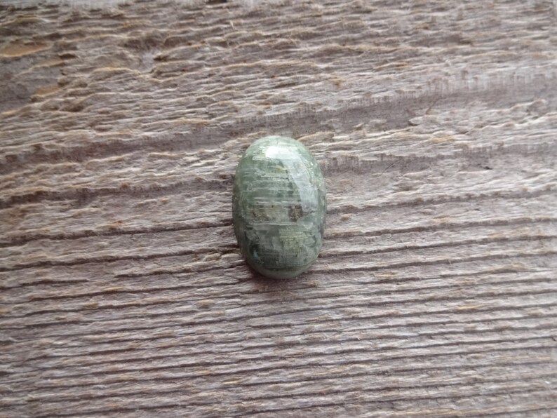K&ouml;nnte beinhalten: Ein gr&uuml;ner ovaler Cabochon-Edelstein mit einem einzigartigen Muster aus wei&szlig;en und grauen Streifen. Der Stein ist vor einem rustikalen Holzhintergrund platziert.