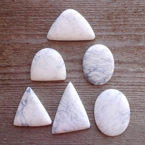May include: Six white and gray cabochon stones in different shapes, including oval, triangle, and teardrop, arranged in a pyramid shape on a wooden surface.