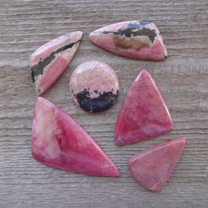 May include: Six pink and black rhodochrosite cabochons of various shapes and sizes. The stones are arranged in a circular pattern on a wooden surface.