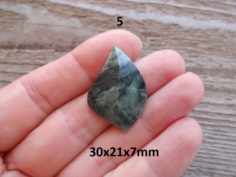 Puede incluir: Un colgante de piedra preciosa verde y negra en forma de l&aacute;grima, que mide 30x21x7mm. El colgante se sostiene en una mano.