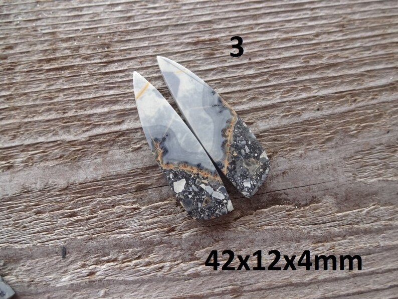 Puede incluir: Dos colgantes de piedra pulida con un patr&oacute;n gris, blanco y negro, y un toque de naranja. Los colgantes tienen forma de tri&aacute;ngulos alargados. El texto "3" est&aacute; encima de los colgantes y "42x12x4mm" debajo. Se muestran sobre una superficie de madera.