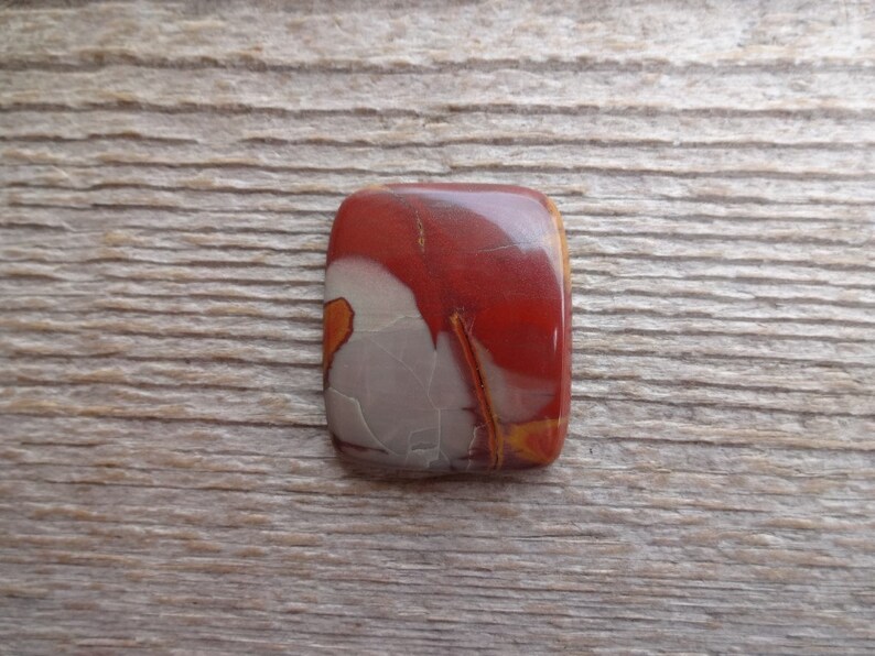 May include: A red, white, and orange cabochon gemstone with a unique pattern. The gemstone is rectangular and has a smooth, polished surface.
