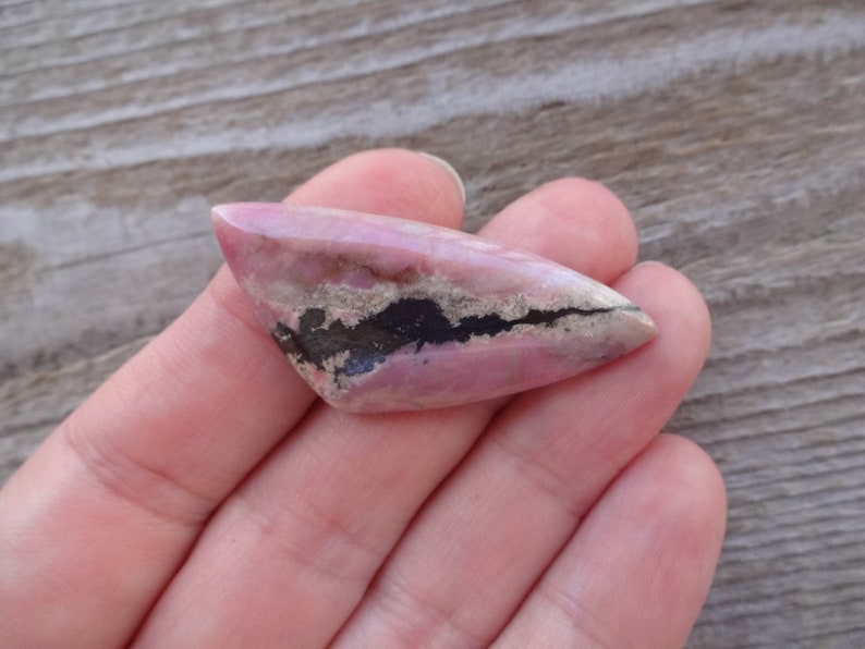 May include: A pink and black cabochon gemstone with a pointed shape. The gemstone has a black band running through the center.