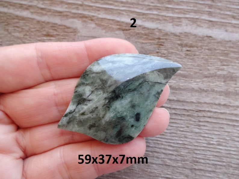 Puede incluir: Una piedra verde y negra con forma de hoja con una superficie lisa y pulida. La piedra mide 59x37x7mm.