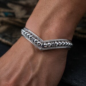 May include: A silver bracelet with a V-shaped design. The bracelet features a series of small, round beads and intricate detailing. The bracelet is worn on a wrist, showcasing its design and craftsmanship.