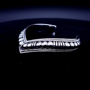 May include: A silver cuff bracelet with a V-shaped design. The bracelet features a row of small, round beads and a twisted rope-like pattern along the edges. The open cuff design allows for easy wear.