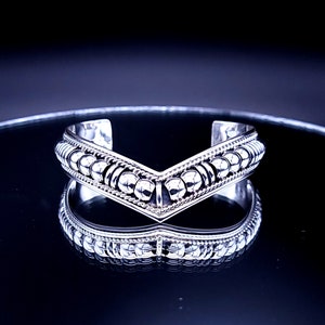 May include: A silver cuff bracelet with a V-shaped design. The bracelet features a row of small, round beads along the top and bottom edges, with a textured pattern in between. The bracelet is reflected on a shiny surface.