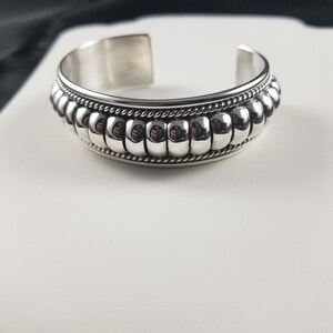 Op de afbeelding: Een zilveren armband met een kralenontwerp. De kralen zijn in een rij rond de armband gerangschikt en worden gescheiden door een touwachtig ontwerp.