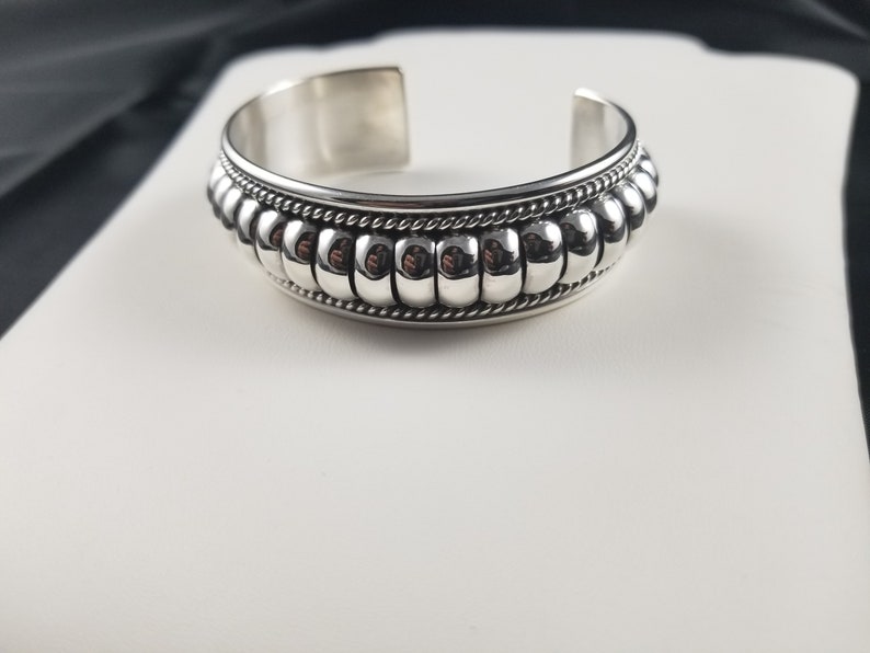 Op de afbeelding: Een zilveren armband met een kralenontwerp. De kralen zijn in een rij rond de armband gerangschikt en worden gescheiden door een touwachtig ontwerp.
