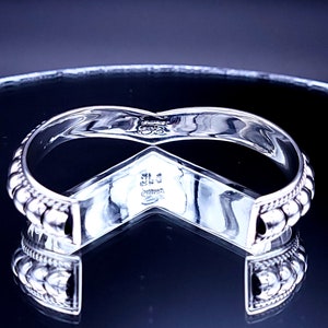 May include: A silver-toned cuff bracelet with a V-shaped design. The bracelet features a beaded edge and a polished finish. The inside of the bracelet has engraved markings. The bracelet is displayed on a reflective surface.