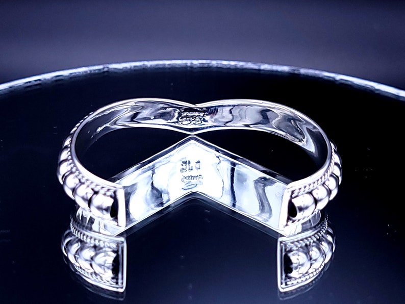 May include: A silver-toned cuff bracelet with a V-shaped design. The bracelet features a beaded edge and a polished finish. The inside of the bracelet has engraved markings. The bracelet is displayed on a reflective surface.