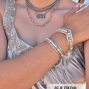 May include: A layered silver necklace and matching bracelets are displayed. The necklace features multiple strands of beads and a decorative pendant. The bracelets have a detailed design. The image includes the text "IG & TikTok: @michelledarlenee".