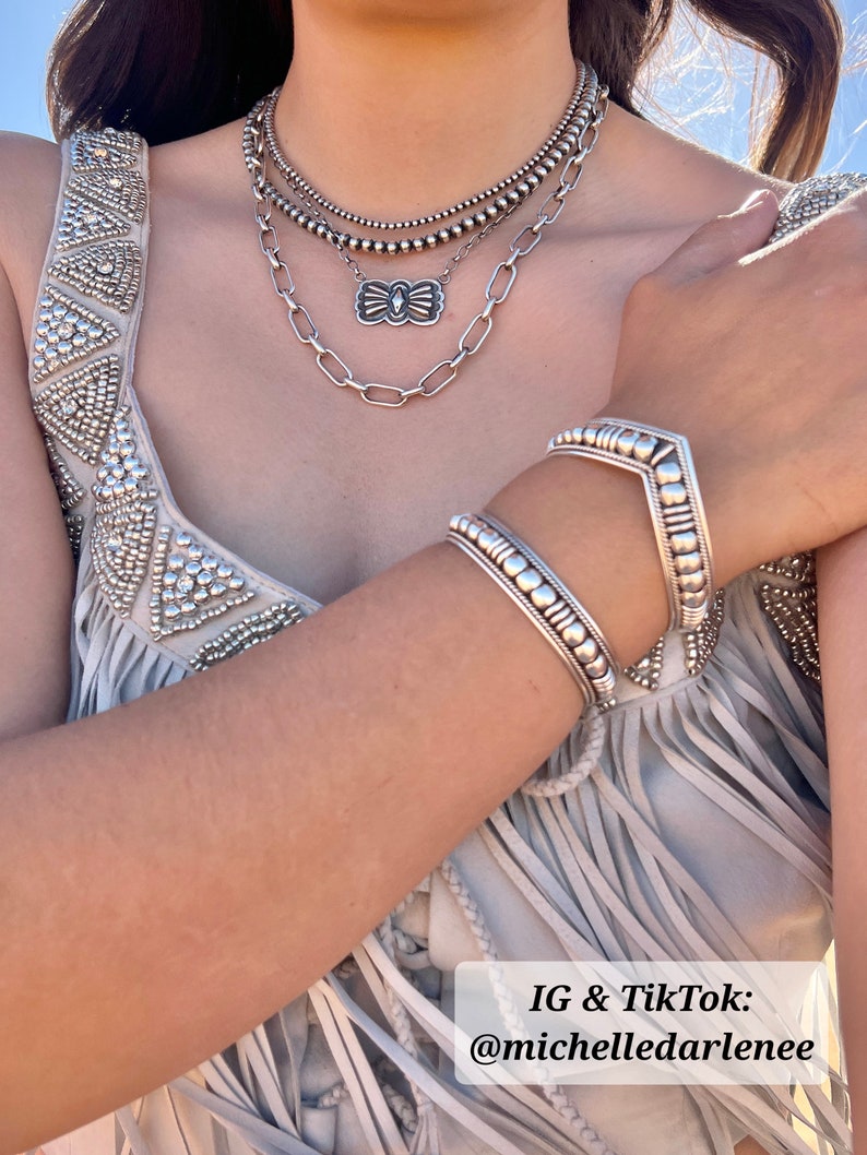 May include: A layered silver necklace and matching bracelets are displayed. The necklace features multiple strands of beads and a decorative pendant. The bracelets have a detailed design. The image includes the text "IG & TikTok: @michelledarlenee".