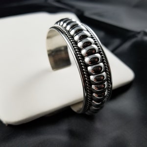Op de afbeelding: Een zilveren armband met een getextureerd, parelmoerachtig ontwerp. De armband is open en heeft een scharniergesp.