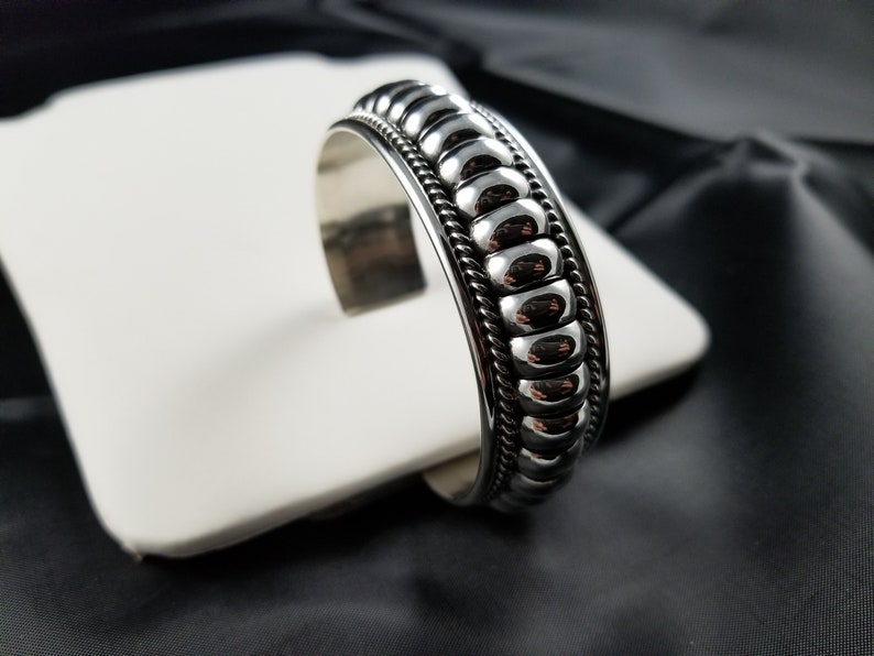 Op de afbeelding: Een zilveren armband met een getextureerd, parelmoerachtig ontwerp. De armband is open en heeft een scharniergesp.