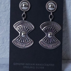 May include: A pair of sterling silver earrings with a detailed design. Each earring features a circular top with a rope-like border, connected to a stylized fan-shaped pendant with intricate patterns and a central sphere. The earrings are displayed on a black card.